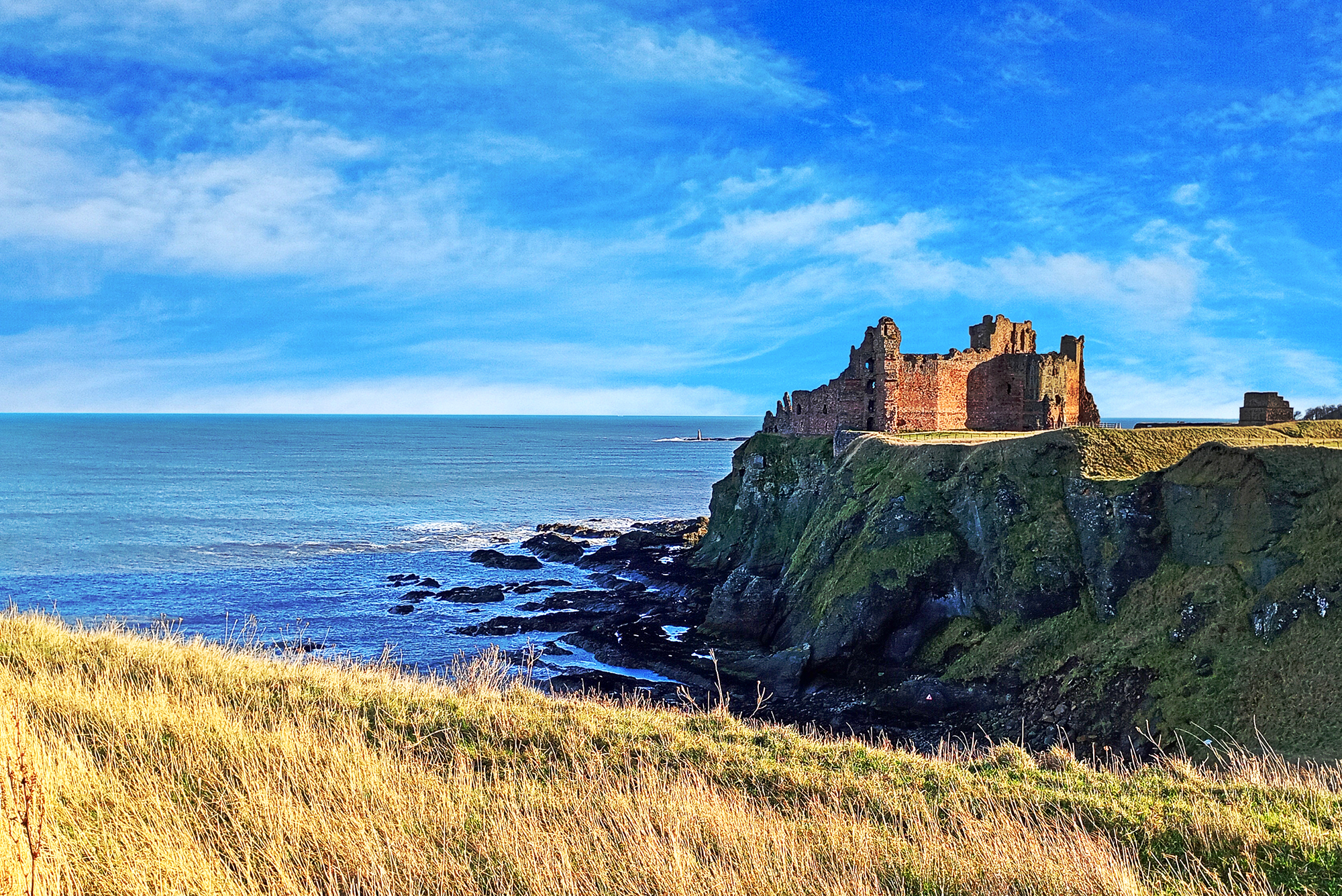 Tantallon Castle with grass in the foreground and the sea and a bright blue sky in the background.