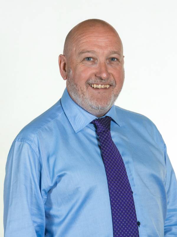 Colin McGinn smiles at the camera, wearing a blue shirt and purple patterned tie.