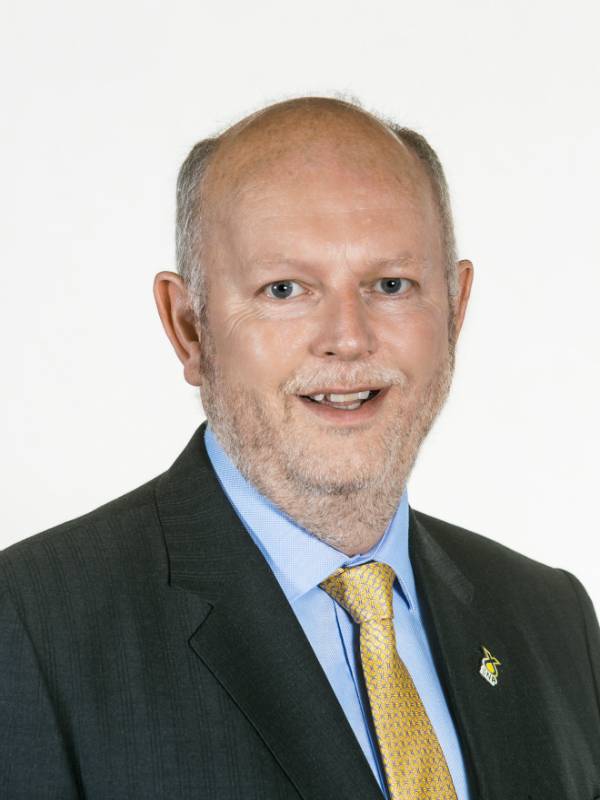 Neil Gilbert smiles at the camera. He is wearing a dark grey suit jacket, a blue shirt and yellow tie. He has a gold brooch on his lapel.