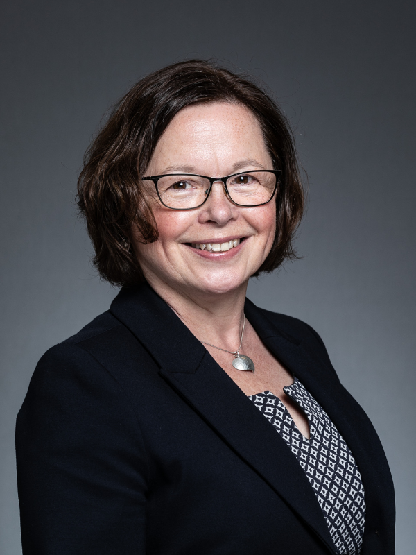 Fiona Dugdale sits side on to the camera, smiling with her face turned towards it. She is wearing a black blazer and a black and white patterned top.