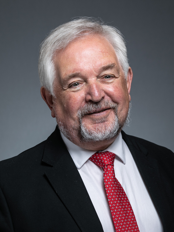 John McMillan smiles past the camera at the photographer. He is wearing a black suit jacket, red patterned tie and white shirt.