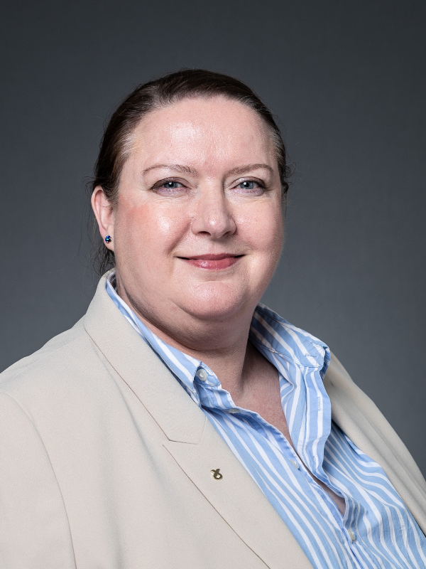 Councillor Liz Allan sits side on with her face turned to the camera. She is smiling, wearing a beige blazer and white and blue striped shirt.