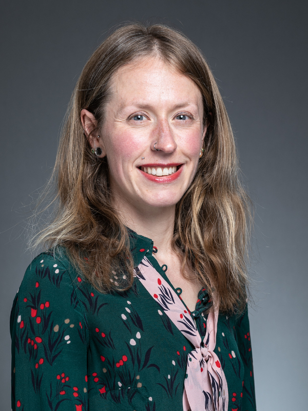 Shona McIntosh smiles at the camera. She is wearing a green floral dress with a matching pink scarf.