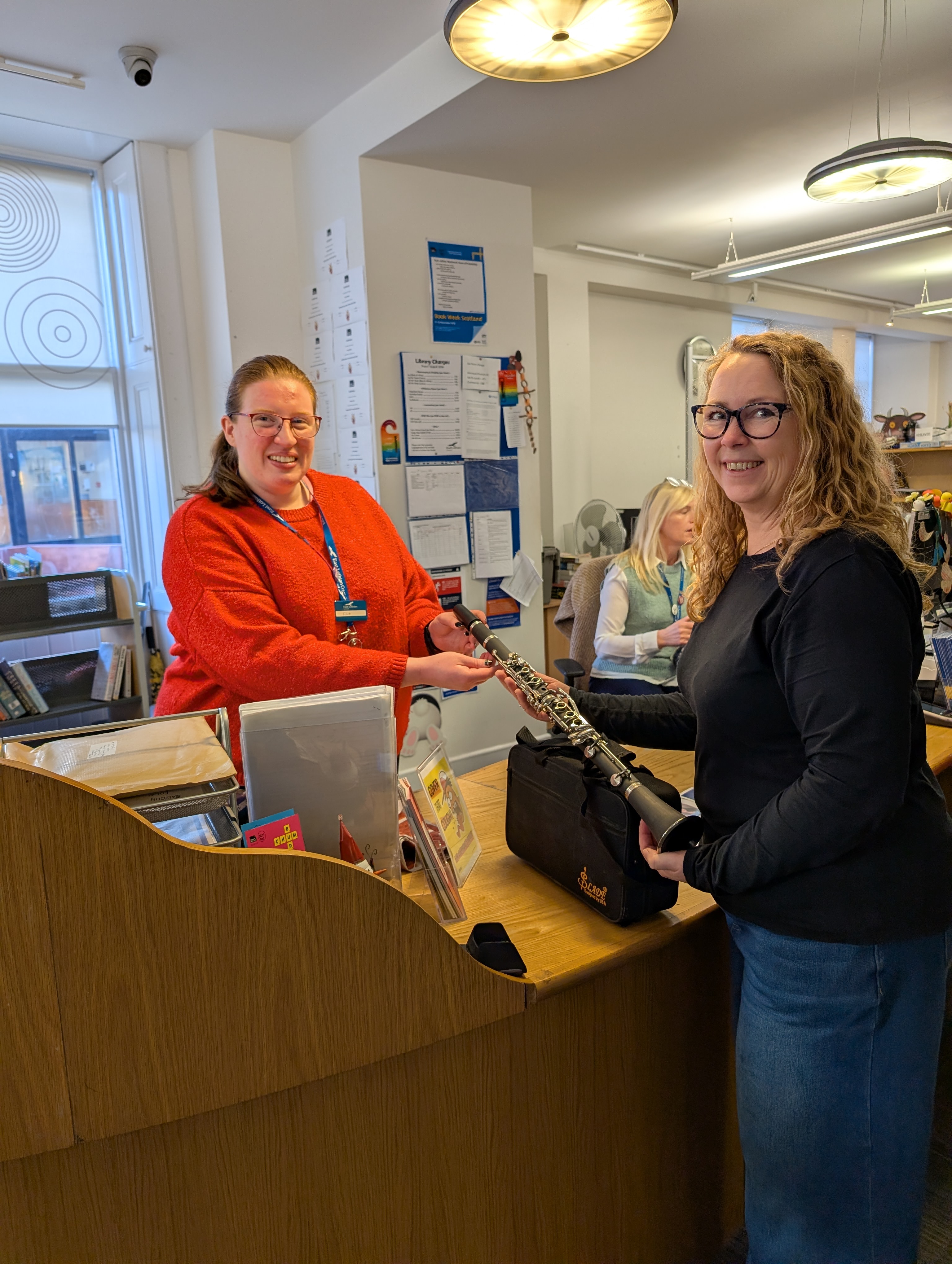 Eilidh wearing a red jumper is standing behind a library desk accepting a clarinet from Mona. Both women are smiling. 