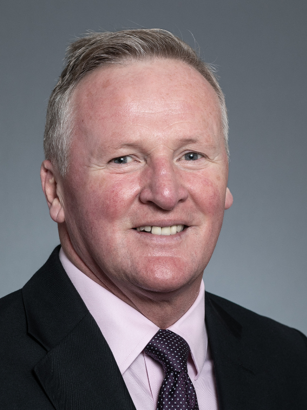 Kenny McLeod smiles at the camera. He is wearing a black suit jacket and tie with a pink shirt.