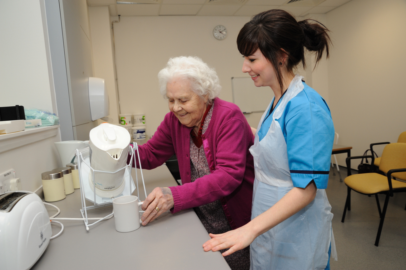 therapist is showing an older woman how to use an adaptive kettle support
