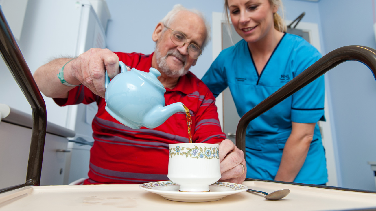Older man is pouring a cup of tea with cup on an assistance trolley
