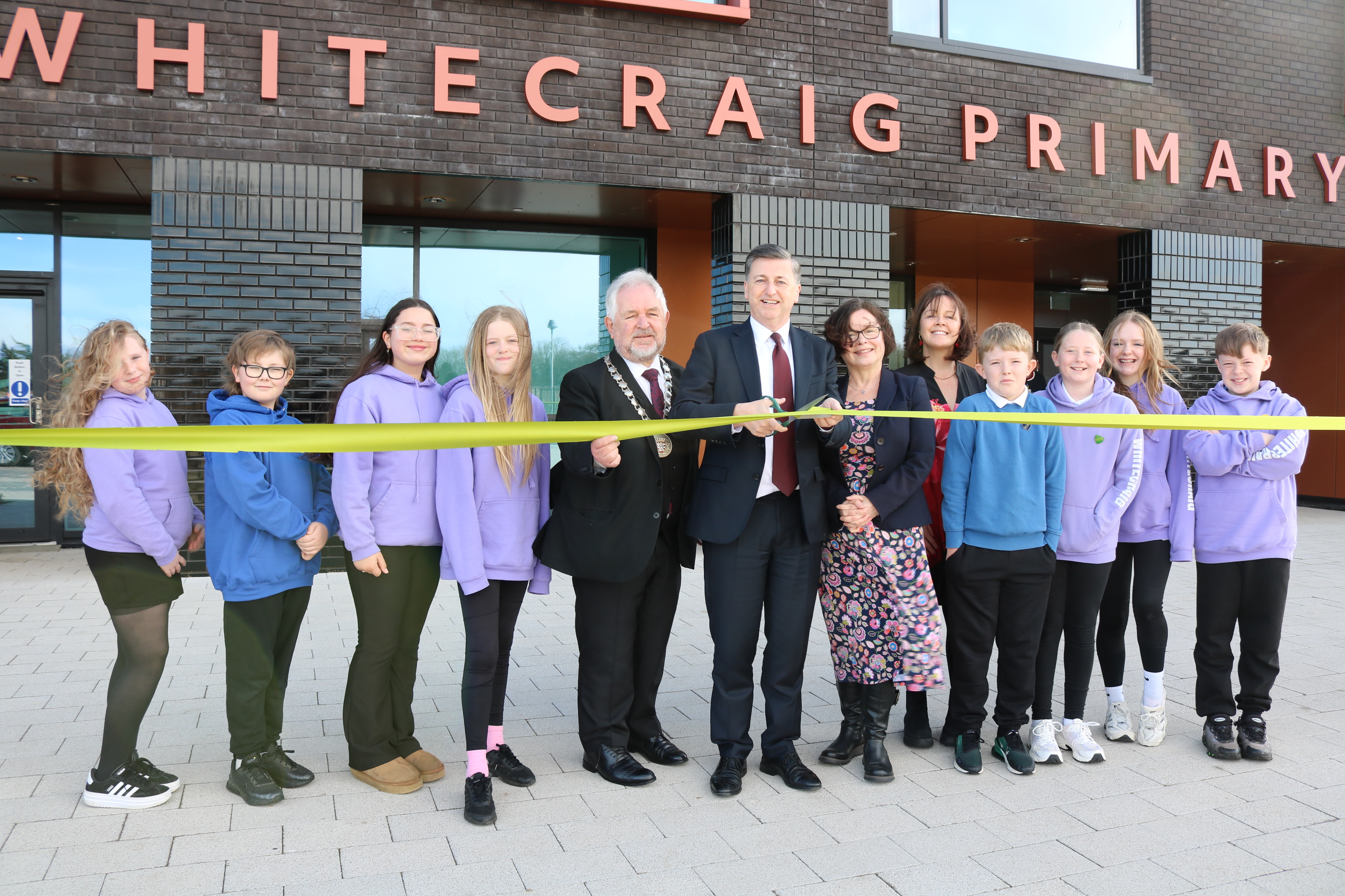 A group of children and adults stand outside Whitecraig Primary School in front of a ribbon