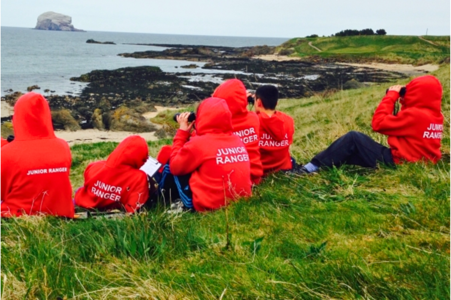 A group of Junior Rangers with binoculars wildlife watching on a cliff with the bass Rock in the background.