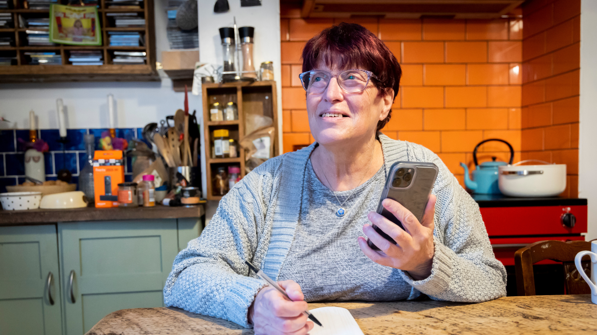 woman sat at kitchen table writing notes while holding a mobile phone