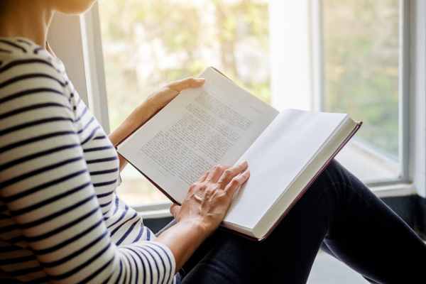 A woman in a stripy top and dark trousers sitting by an open window reading a book. There are trees outside the window.